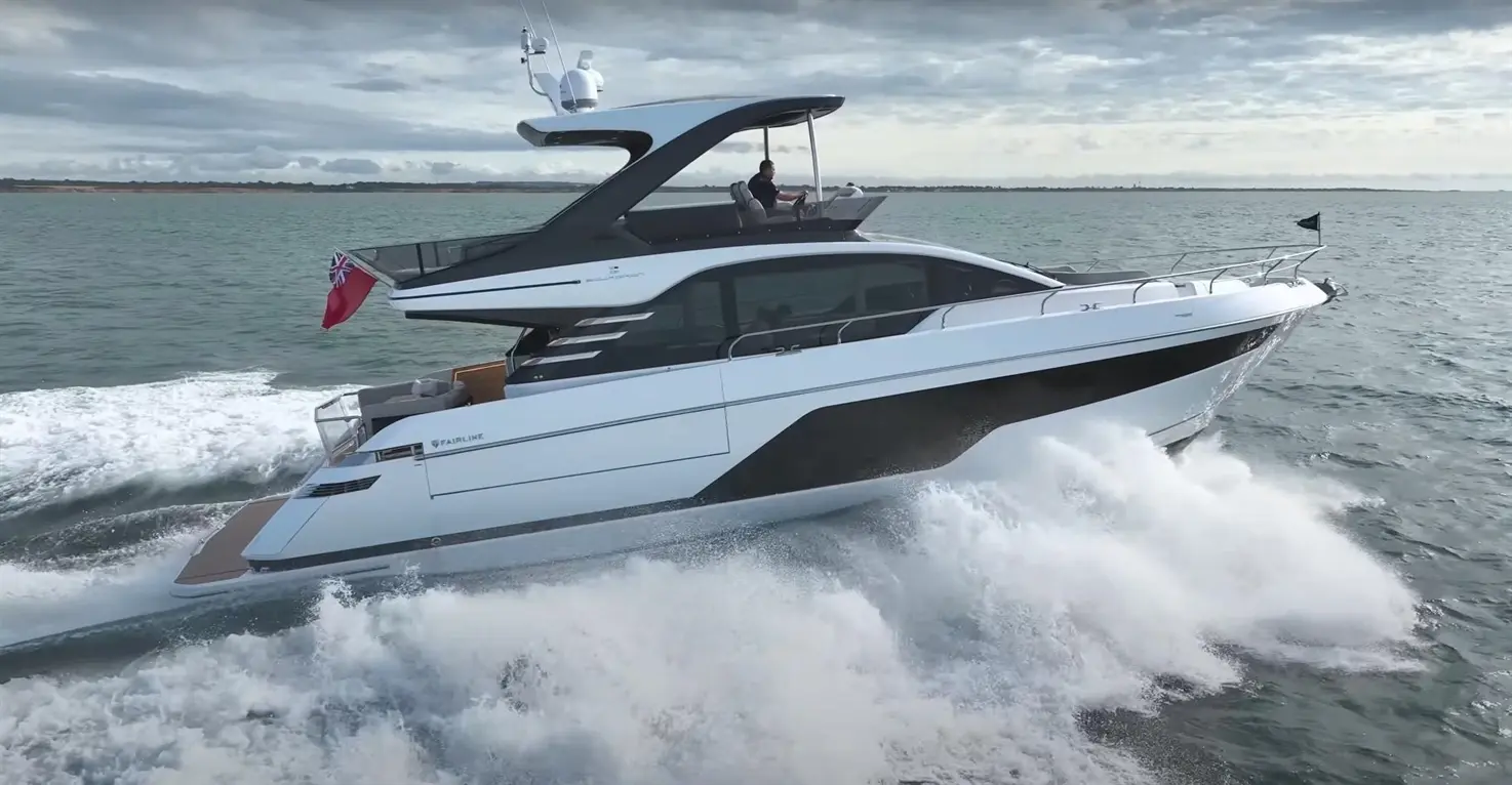 A sleek Fairline yacht speeds across water, leaving a spray of waves, with a captain at the helm and a flag flying.
